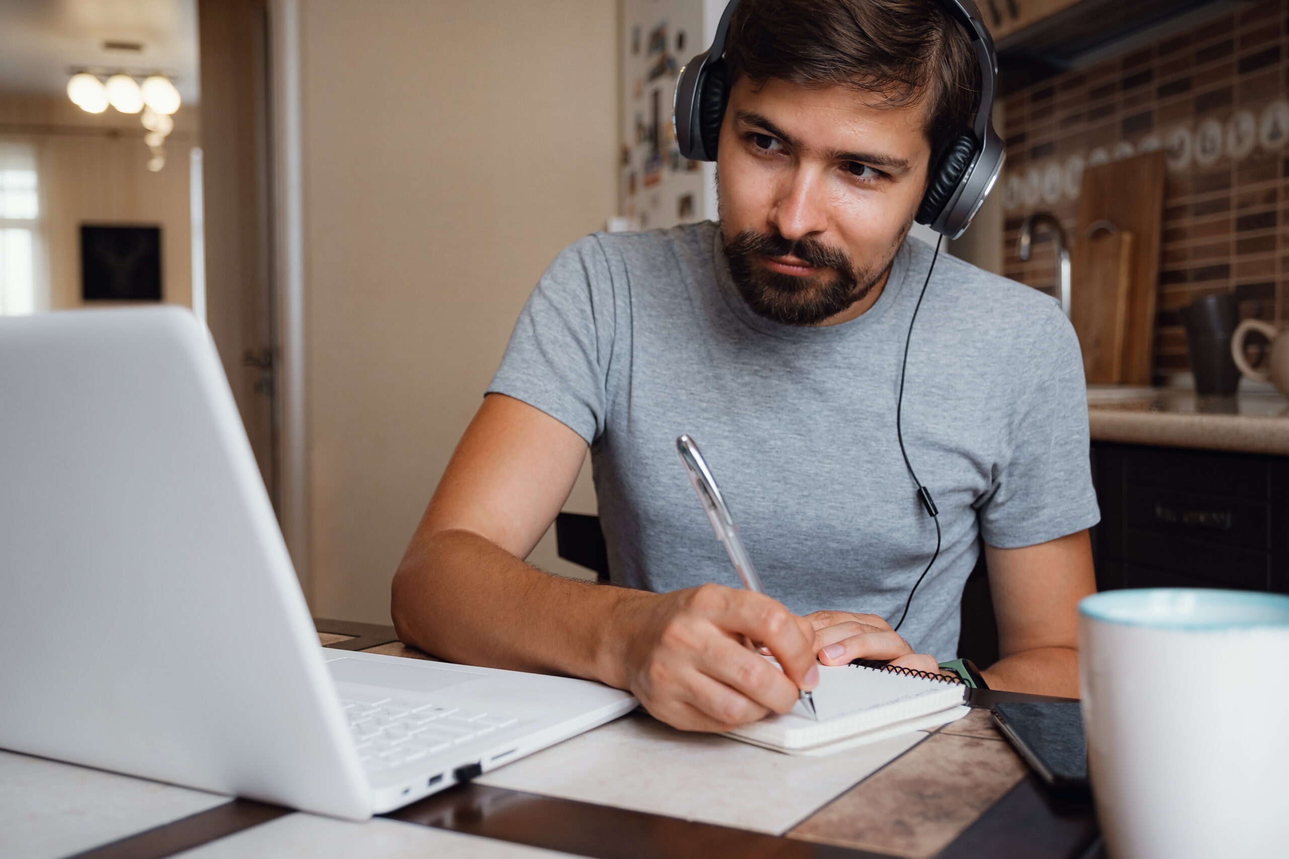Focused young man wear headphones study online watching webinar podcast on laptop listening learning education course conference calling make notes sit at work desk, elearning concept