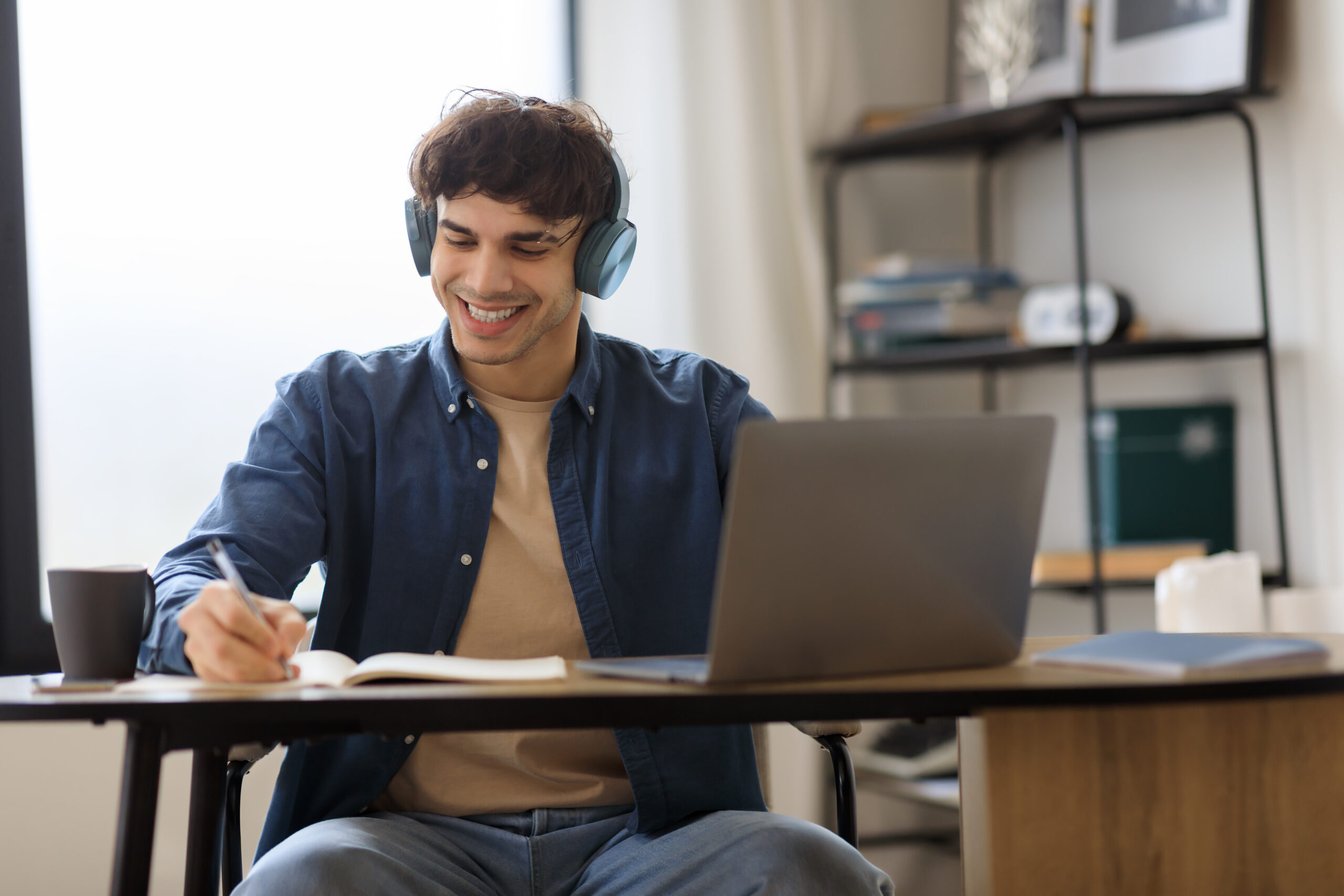 Successful Middle Eastern Entrepreneur Man At Laptop Computer Taking Notes Wearing Earphones, Learning And Working Online Sitting At Desk Indoor. E-Learning Leisure Concept